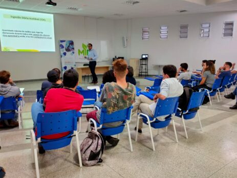 Grupo de estudantes sentados em um auditório enquanto participam de uma palestra com apresentação de slides em projetor.