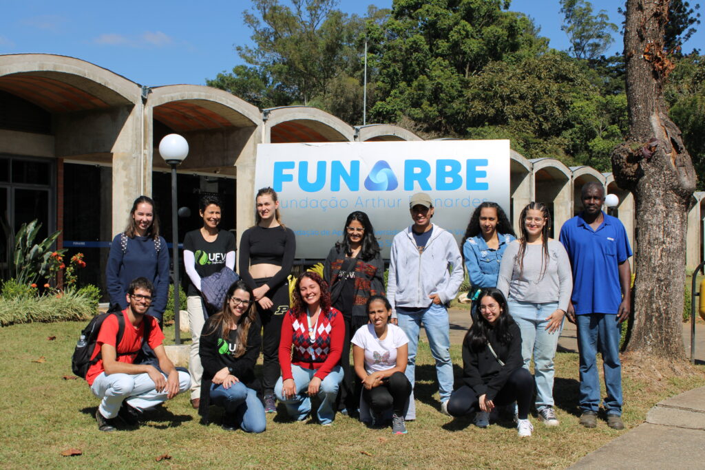 Estudantes nacionais e estrangeiros da UFV ao lado de colaboradores da Funarbe, posando para foto em frente à placa com logotipo da fundação.