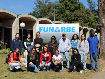 Estudantes nacionais e estrangeiros da UFV ao lado de colaboradores da Funarbe, posando para foto em frente à placa com logotipo da fundação.